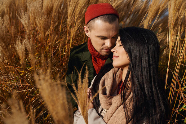 A couple wrapped in warm attire share a tender moment in a field of autumn grass.