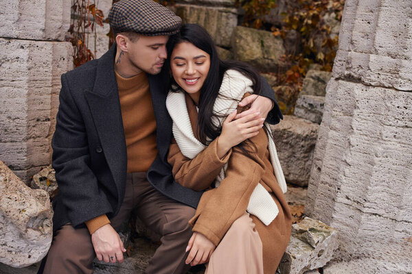 A warm embrace between a couple dressed for autumn amid rustic stone ruins and vibrant foliage.