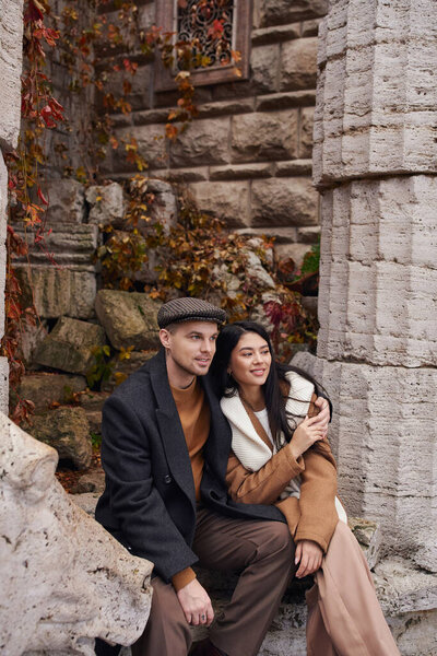 A couple embraces each other warmly, surrounded by vibrant autumn foliage and weathered stone.