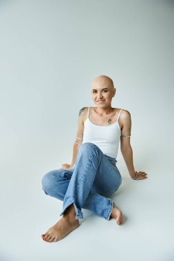 Saçları dökülmüş genç bir kadın rahat bir şekilde pürüzsüz bir yüzeyde oturur, özgüven ve zarafet saçar..