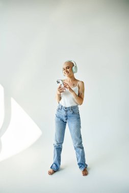 A young bald woman looks at her phone, immersed in music, in a minimalist studio setting. clipart