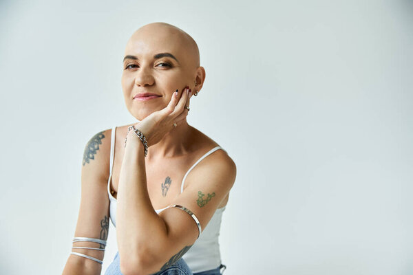 A young bald woman confidently poses with a thoughtful expression, showcasing her tattoos.
