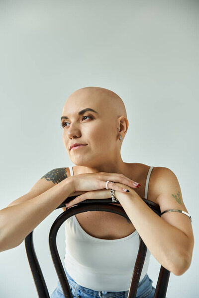 A young bald woman smiles softly while resting her arms on a chair, radiating confidence.