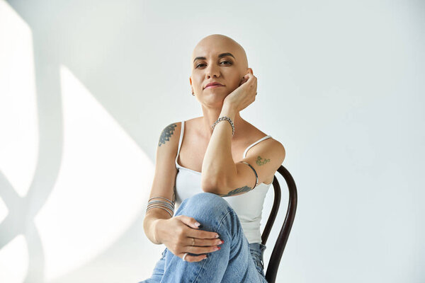 Молодая женщина с алопецией задумчиво сидит на стуле, демонстрируя свою уверенность и изящество..