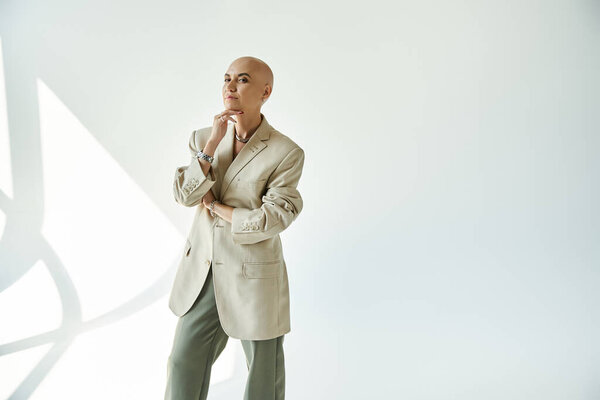 Уверенная молодая лысая женщина задумчиво позирует в шикарной одежде, демонстрируя изящество и силу.