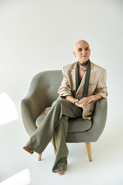 Молодая женщина с алопецией элегантно сидит в кресле, излучая уверенность.