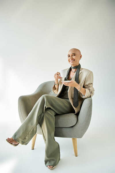 Молодая лысая женщина с алопецией элегантно сидит в уютном кресле, наслаждаясь чаем..