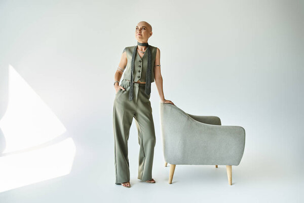 Молодая лысая женщина уверенно стоит рядом с современным стулом, демонстрируя свою неповторимую красоту.