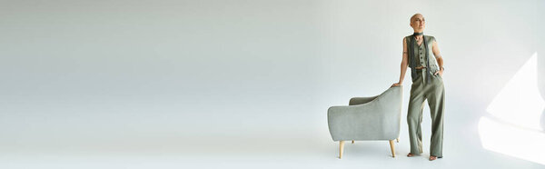 Молодая лысая женщина уверенно позирует рядом со стильным стулом в спокойной обстановке.