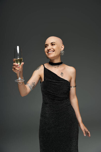 Сияющая молодая лысая женщина празднует в элегантном черном платье, радостно поднимая бокал.
