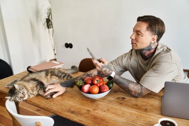 A young man engages with his phone while lounging at home beside his relaxed cat and fresh produce. clipart