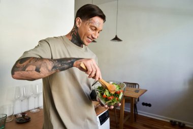 Genç yakışıklı bir adam, evdeki rahat mutfağında canlı bir salata hazırlarken gülümsüyor..