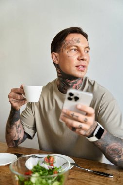 Genç bir adam kahvesini yudumluyor ve akıllı telefonunu kontrol ediyor, evde taze salata ile çevrili..