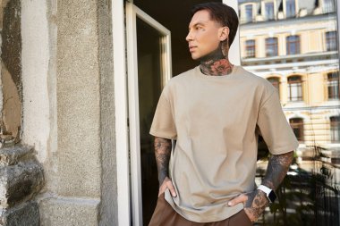 A stylish young man with tattoos gazes thoughtfully outside from his home balcony. clipart