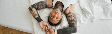 Yakışıklı genç bir adam yatakta uzanıyor, çarpıcı dövmelerini ve rahat tavırlarını sergiliyor..
