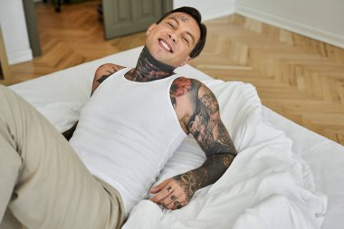 Genç adam bir yatakta uzanır, dövmeleri görünür ve sıradan bir stil ile neşe saçar..