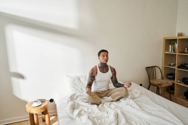 Yakışıklı genç bir adam yatakta bacak bacak üstüne atarak oturur. Parlak bir odada huzur içinde meditasyon yapar..
