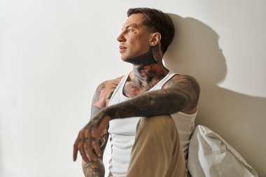 Dövmeli genç bir adam düşünceli bir şekilde evinde oturuyor, sükunetin ve doğal ışığın tadını çıkarıyor..