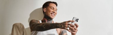 A young, tattooed man enjoys a lighthearted moment while using his smartphone at home. clipart