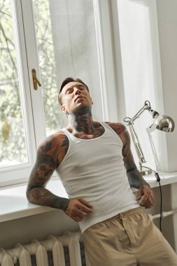 A young, tattooed man leans back, soaking in the warmth of sunlight while at home. clipart