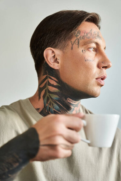 A young man sips coffee, revealing intricate tattoos in a serene setting.