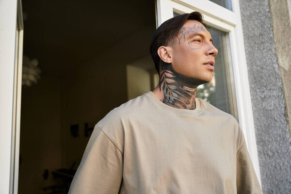 Standing at the doorway, a tattooed young man contemplates the world outside his home.