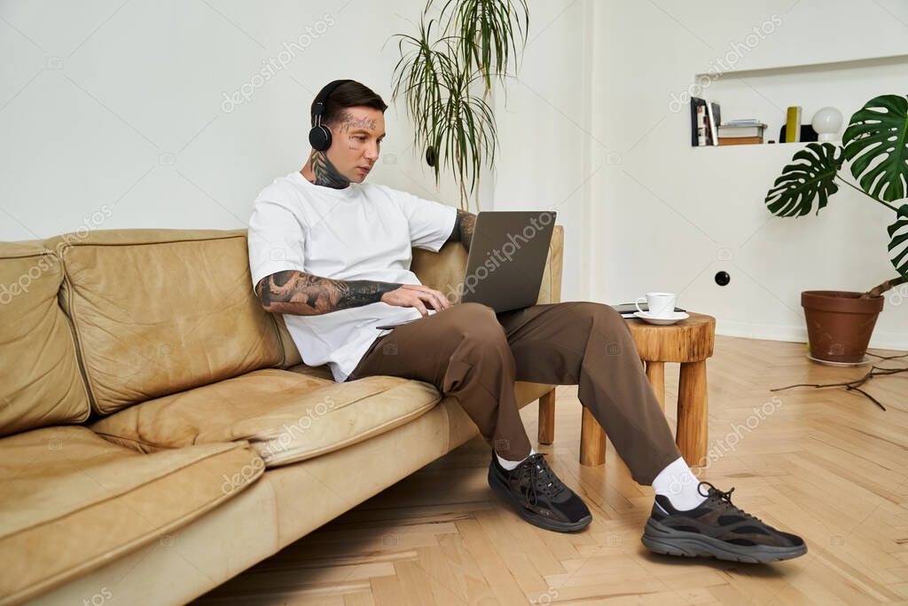 A young man sits comfortably on a couch, focused on his laptop while enjoying music and coffee.
