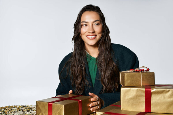 Изысканная молодая женщина тепло улыбается, обнимая праздничную атмосферу с подарками поблизости.