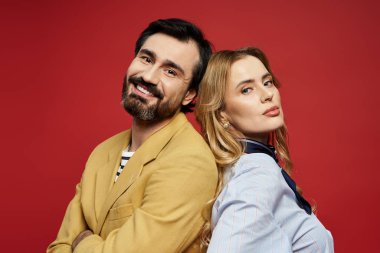 Sarı ceketli orta yaşlı bir adam ve mavi gömlekli bir kadın sırt sırta verip gülümsüyor..