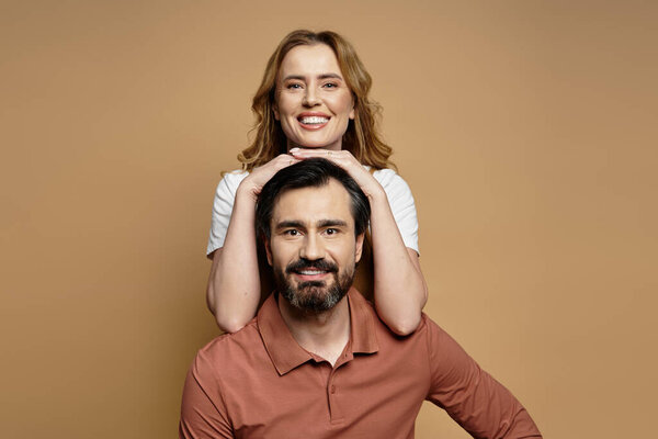 Женщина средних лет игриво позирует позади мужчины, широко улыбаясь и излучая счастье..