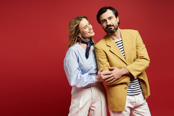 A middle aged couple shares a lighthearted moment against a vibrant red backdrop, smiling joyfully.