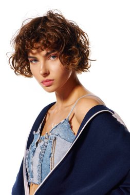 Genç bir kadın, parlak ışık altında kıvırcık saçlarını ve modaya uygun kıyafetlerini sergileyerek düşünceli bir şekilde bakıyor..