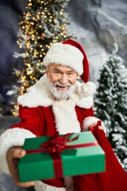 Şenlikli bir Noel Baba, yeşil bir hediye sunar. Ağaçların arasında kırmızı bir yay ile..