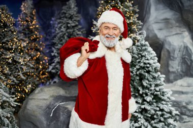 Neşeli bir Noel Baba, karlı bir arazide bir hediye çantası tutuyor..