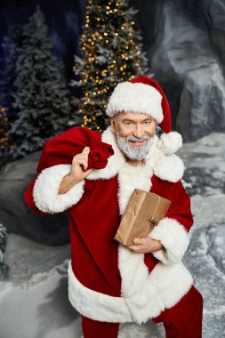 Noel Baba, karlı bir arazide paketlenmiş bir hediyeyi tutarken neşeli neşesini paylaşıyor..
