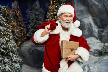 Neşeli bir Noel Baba, elinde bir hediye çantası ve neşeyle karlı ağaçların arasında duruyor..