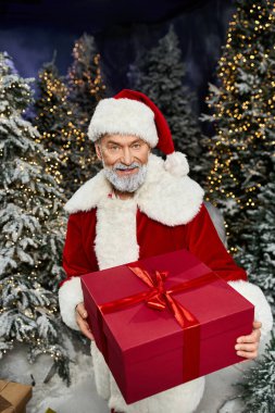 Noel Baba 'nın elinde karla kaplı ağaçlar ve parıldayan ışıklarla çevrili büyük kırmızı bir hediye var..