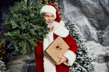 Noel Baba, etrafı karla kaplı ağaçlar ve dekorasyonlarla çevriliyken neşeli bir hediye tutuyor.