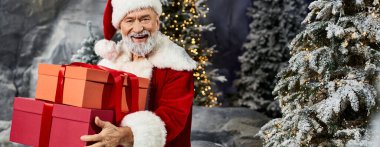 Neşeli bir Noel Baba karla kaplı ağaçlarla çevrili canlı hediyeler tutarken ışıldıyor..