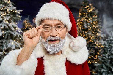 Neşeli bir Noel Baba karlarla kaplı ağaçların arasında duruyor..