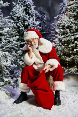 Neşeli bir Noel Baba her zaman yeşil ağaçların arasında oturur ve büyük hediye çuvalının yanında düşünür..
