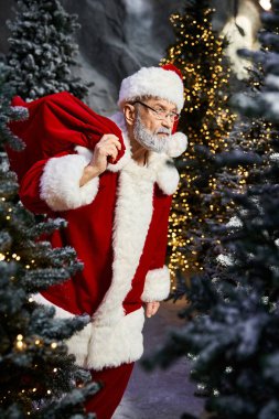 Kırmızı elbiseli neşeli bir Noel Baba etrafı süslenmiş ağaçlarla çevriliyken bir çuval taşıyor..