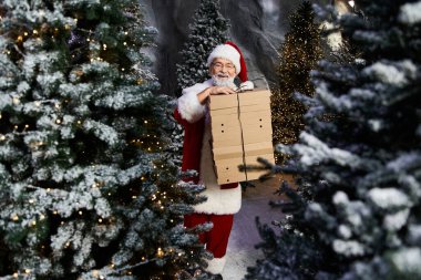 Noel Baba, etrafı karla kaplı Noel ağaçları ve ışıklarıyla çevrili bayram hediyeleri taşıyor..