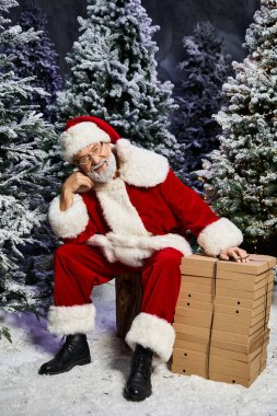 Neşeli bir Noel Baba, kışın harikalar diyarı karla çevrili hediyelerin yanında oturur..