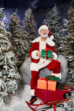 A cheerful figure in red and white attire holds gifts, surrounded by snow dusted trees. clipart