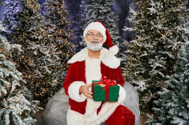 Noel Baba otururken sıcacık gülümser. Etrafı şenlikli ağaçlar ve parıldayan ışıklarla çevrilidir..