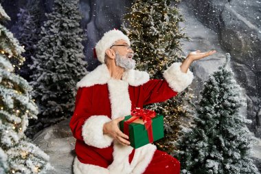 Neşeli bir Noel Baba, etrafı karla kaplı ağaçlarla çevrili bir hediye tutar..