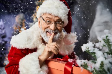 Noel Baba, Noel neşesinin ortasında sarılı bir hediyeyle sessizlik için şakacı jestler yapar..