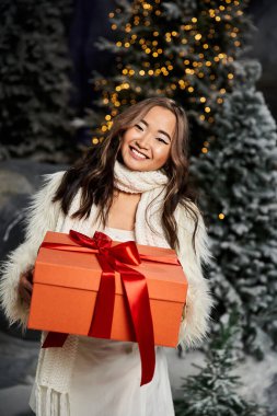 Samimi giyinmiş genç bir kadın Noel süslemeleri arasında bir hediyeyle gülümsüyor..