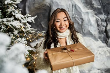 Bir kadın güzel bir Noel arkaplanının ortasında elinde paketlenmiş bir hediyeyle ışıl ışıl gülümsüyor..